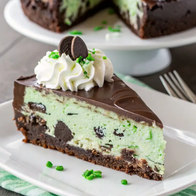 A slice of Thin Mint cheesecake on a white plate with a chocolate ganache topping and crushed Thin Mint cookies on top, photographed from slightly above on a dark wood surface.
